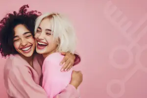 Duas amigas abraçadas, sorrindo com alegria. Uma com cabelo crespo e outra com cabelo loiro e curto, representando diversidade, afeto e autoestima.