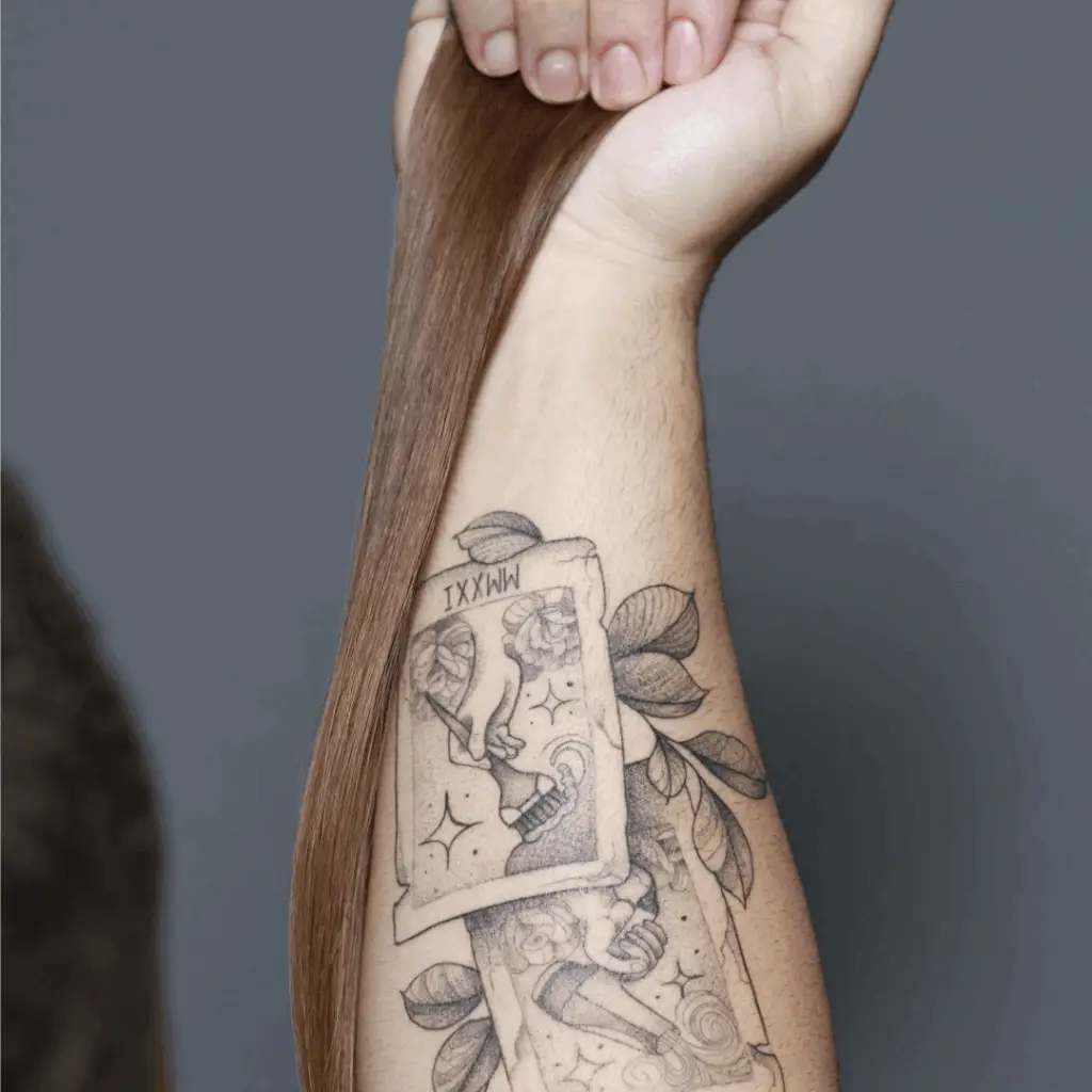 Braço de Davi Moraes com tatuagem de cartas de baralho, cada uma ilustrando ferramentas de cabeleireiro, com uma mecha de extensão capilar repousando sobre o antebraço.