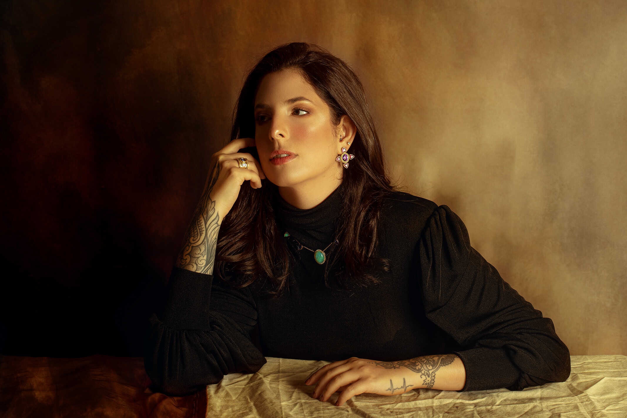 Retrato de Carol Razuk em fundo quente dourado, usando blusa preta, brincos autorais e colar com pedra verde. Ela posa apoiando o rosto na mão, com anéis e tatuagens visíveis.