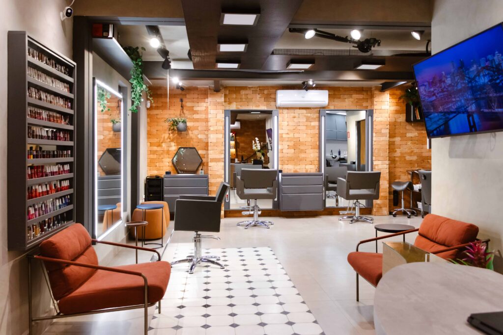 Foto do ambiente principal do Beauty.co.lab, com duas estações de atendimento, espelhos iluminados, parede de tijolos aparentes, poltronas de espera e iluminação técnica. Um espaço desenhado para acolher com calma e permitir execução precisa, alinhado à filosofia de cuidado, comunidade e laboratório.